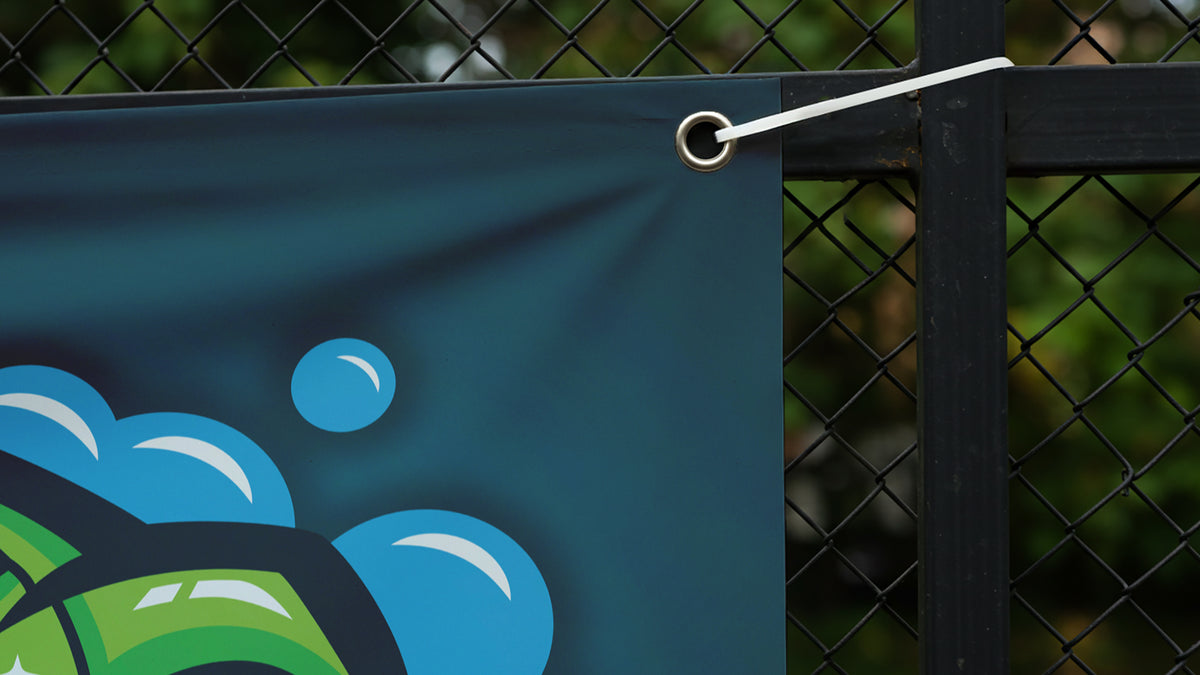 Close-up of a vinyl banner corner, showing the hemmed edge, metal grommet, and zip tie fastening it to a fence