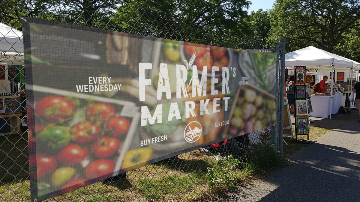Wide mesh banner fastened along a chain-link fence at an outdoor market