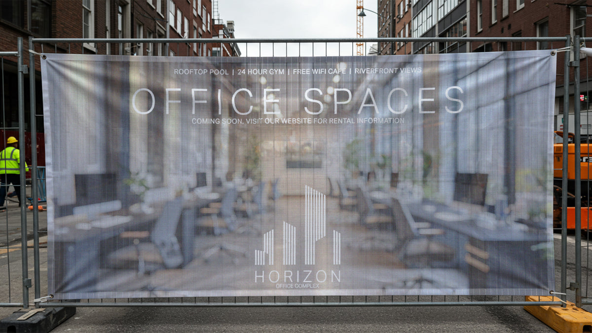 Mesh banner mounted across a construction barrier fence, with the background visible through the weave