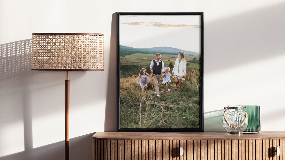 Poster print in a slim black frame, standing on a sideboard beside a lamp